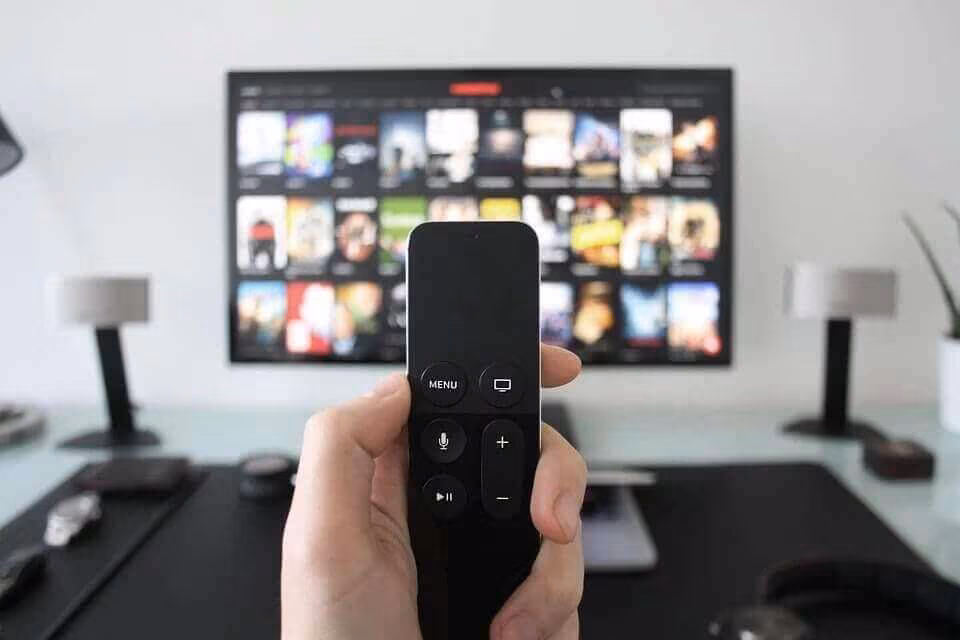 A hand holding a remote control in front of a television showing a streaming menu