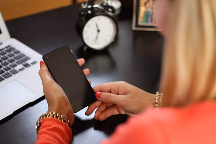 A woman holding a phone at a desk with a laptop and an alarm clock