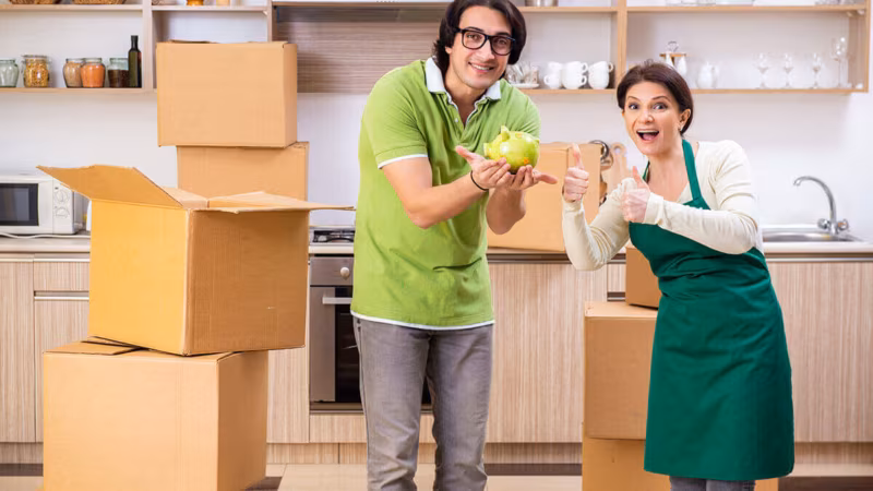 Two people in a kitchen surrounded by cardboard boxes, one holding a plant