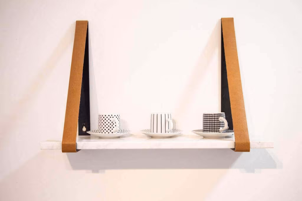 A white shelf suspended from leather straps on a pale wall, holding stacked cups and saucers