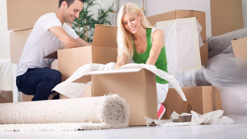 A couple kneeling on the floor to pack a cardboard box, with cushions and a rolled rug beside them