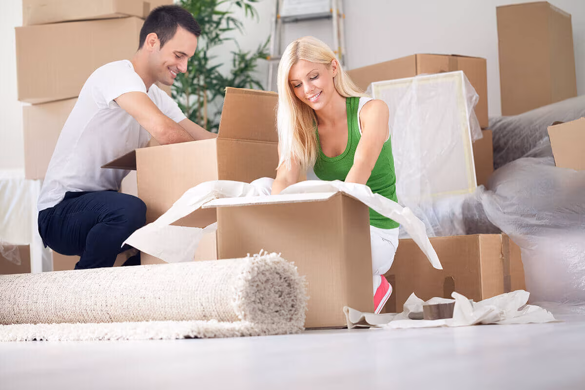 A couple kneeling on the floor to pack a cardboard box, with cushions and a rolled rug beside them