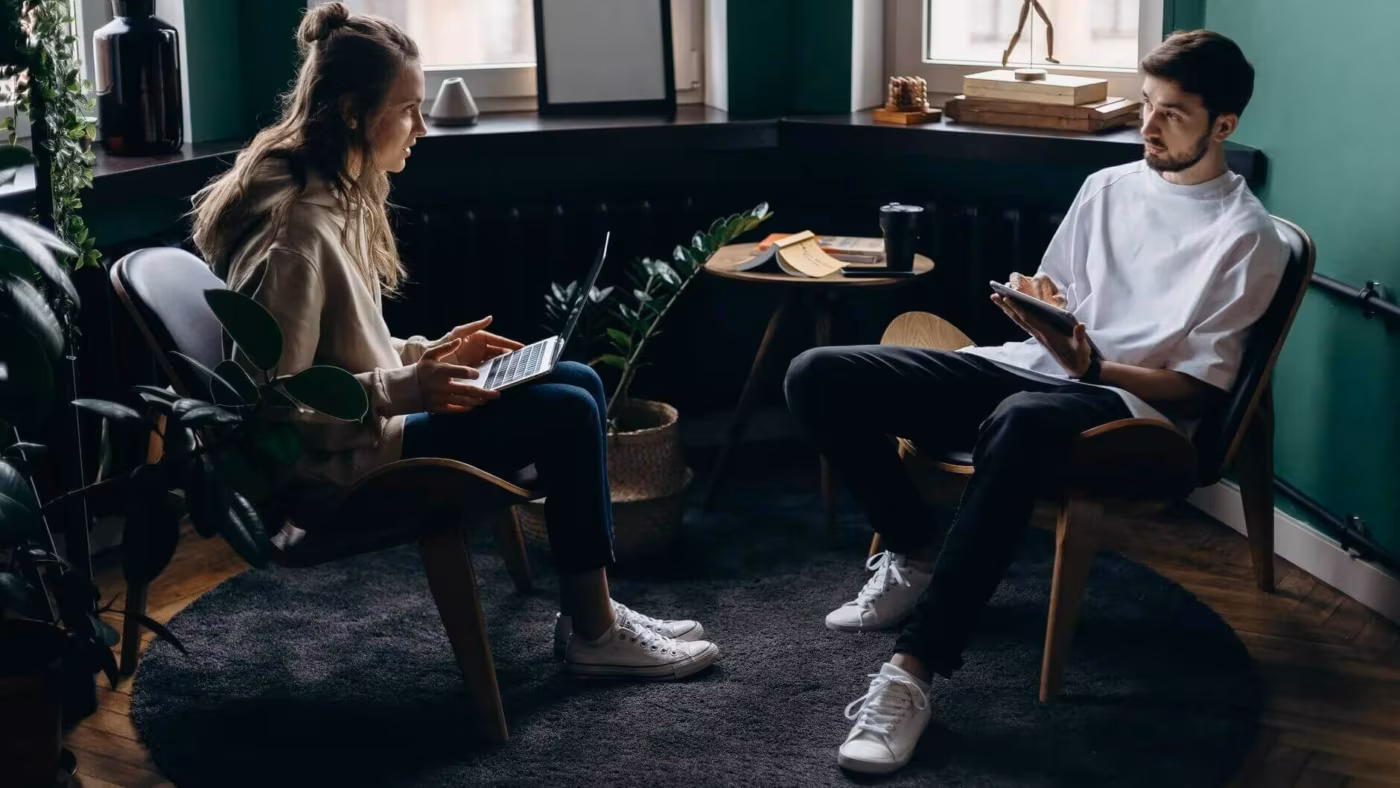 Two people sitting in armchairs in a green-walled lounge, each looking at a device