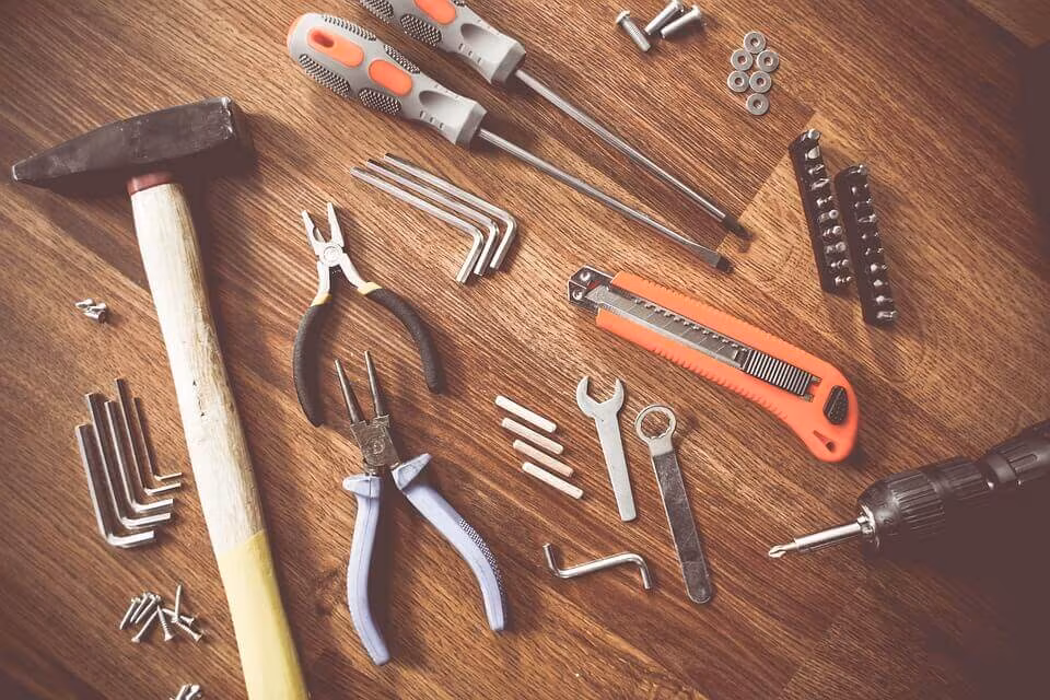 Hand tools laid out on a wooden surface — a hammer, pliers, screwdrivers, spanners and a knife