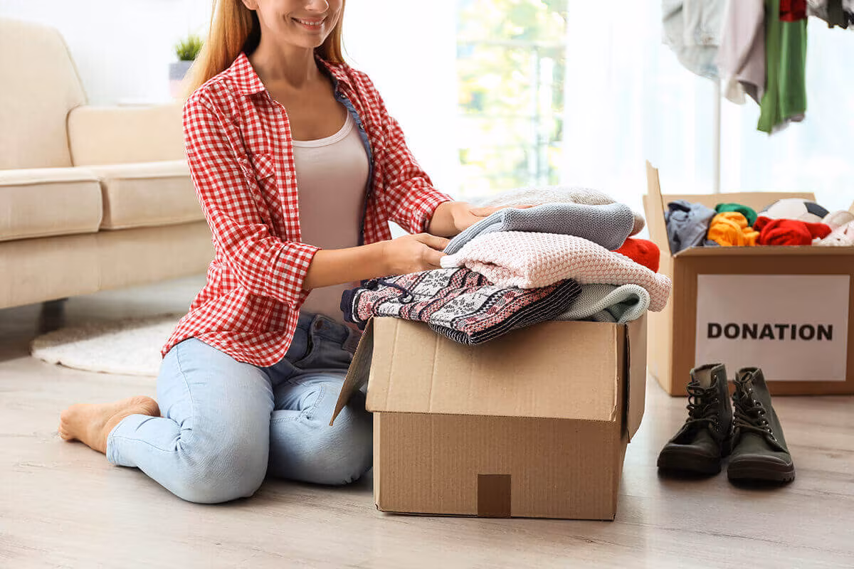 A woman kneeling on the floor folding clothes into a box, with a box marked Donation beside her