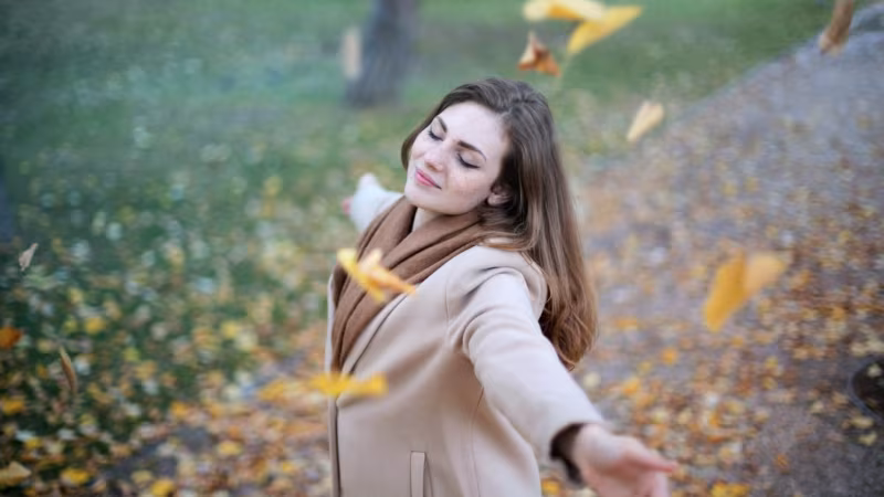 A woman with her arms open and eyes closed among falling autumn leaves