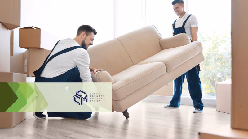 Two movers carrying a beige sofa through a room stacked with boxes