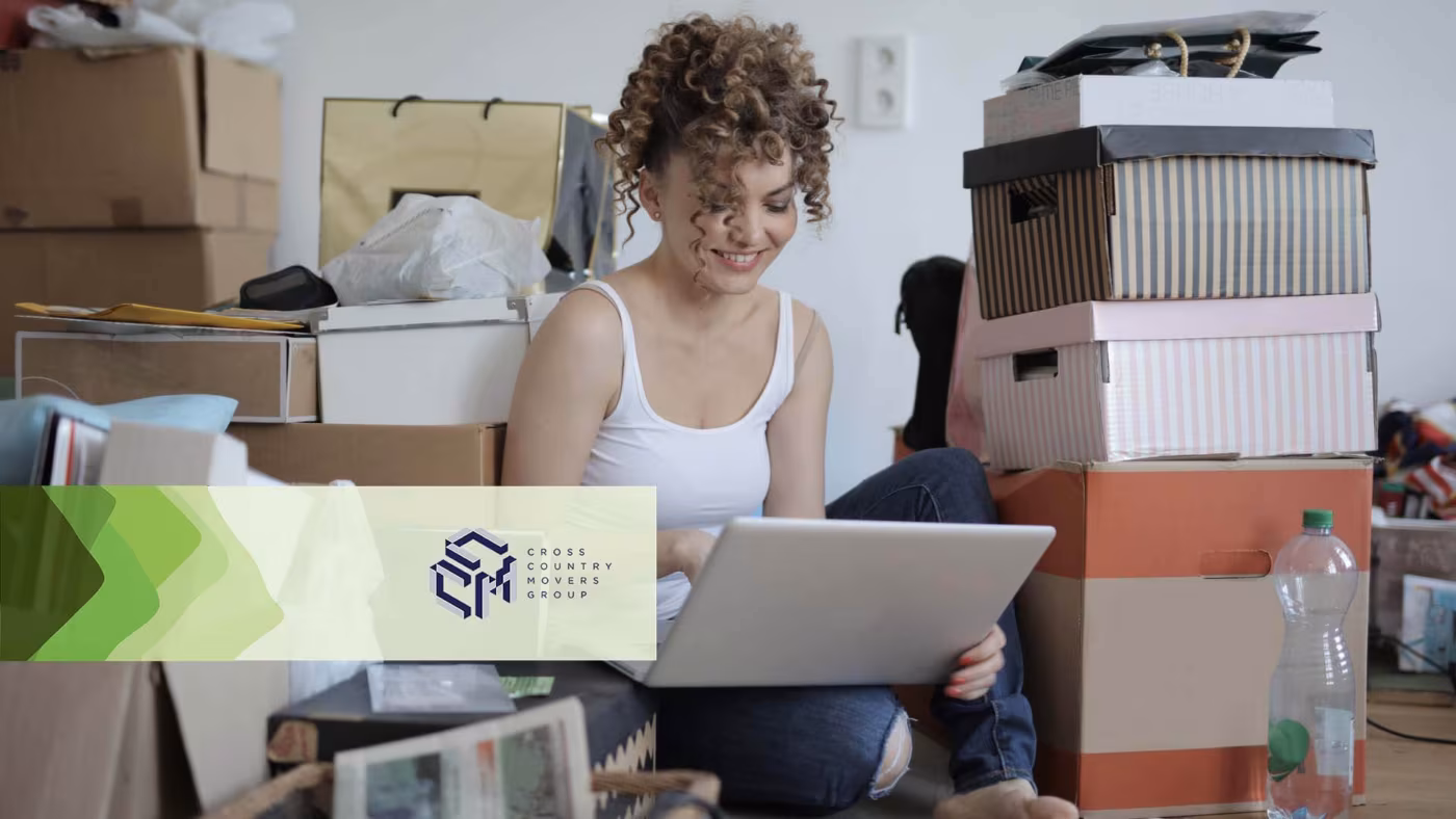 A young woman sitting on the floor with a laptop among cardboard boxes and storage bins
