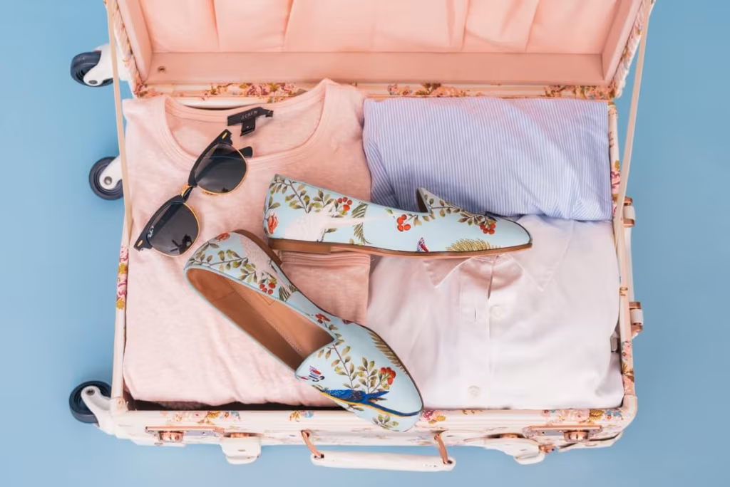 Pink suitcase filled with clothes, a pair of shoes, and sunglasses