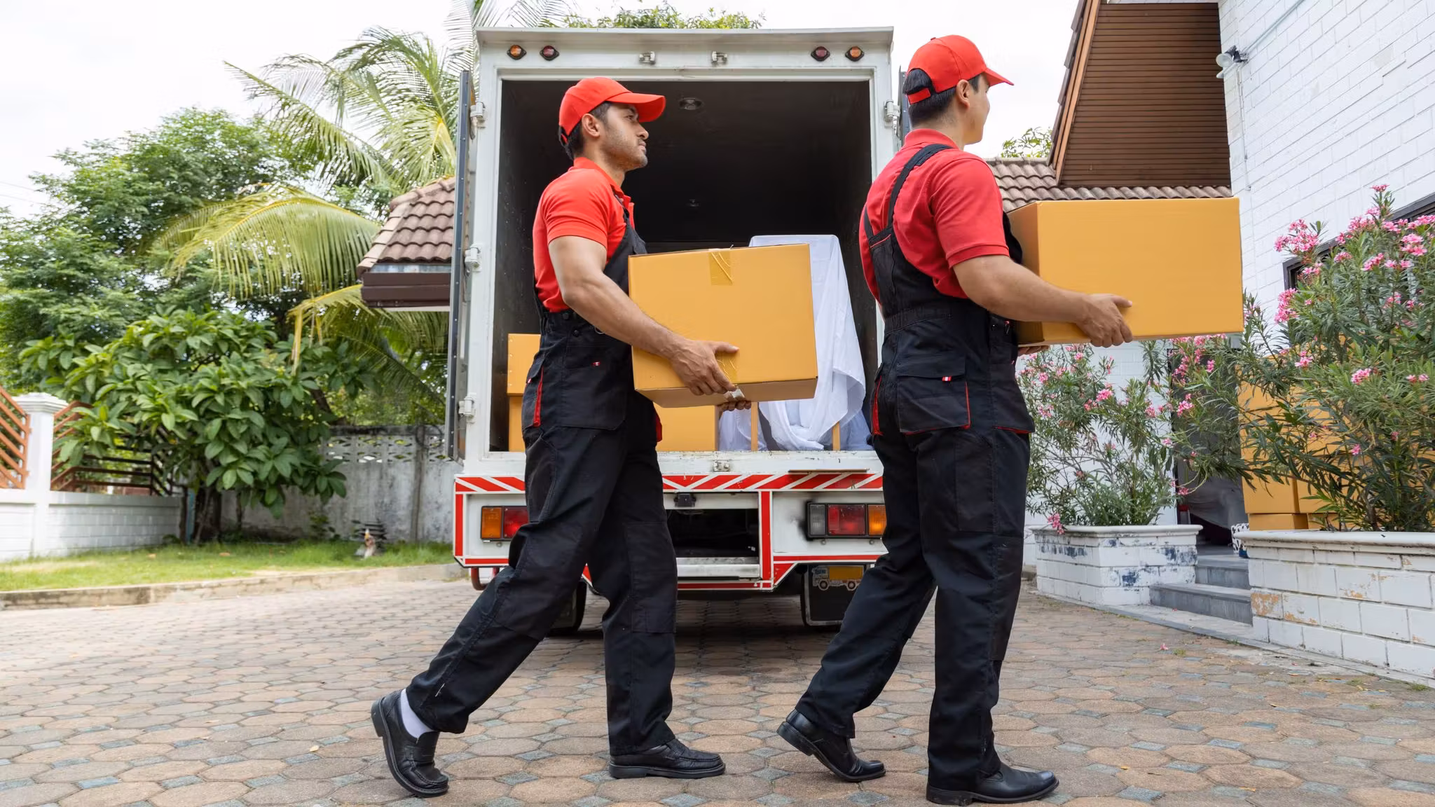 Two uniformed movers carrying cartons up a paved drive from their own open box truck