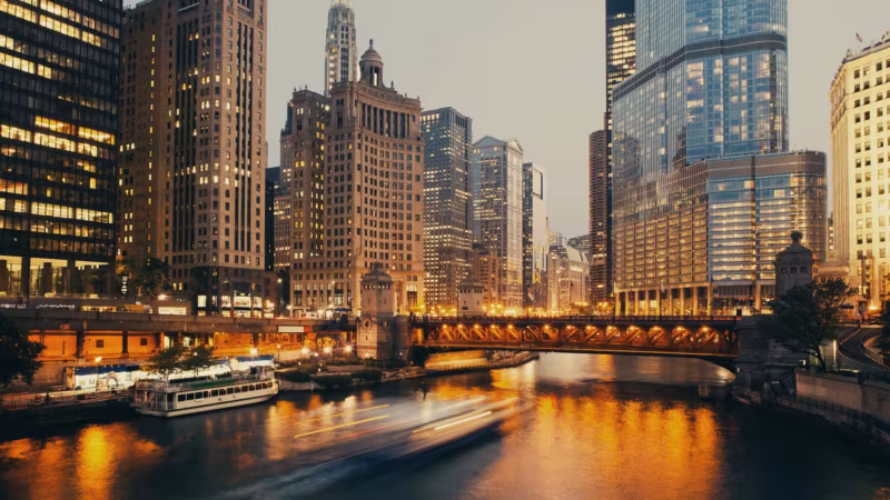 The Chicago River at dusk between downtown towers, a boat passing under a bridge