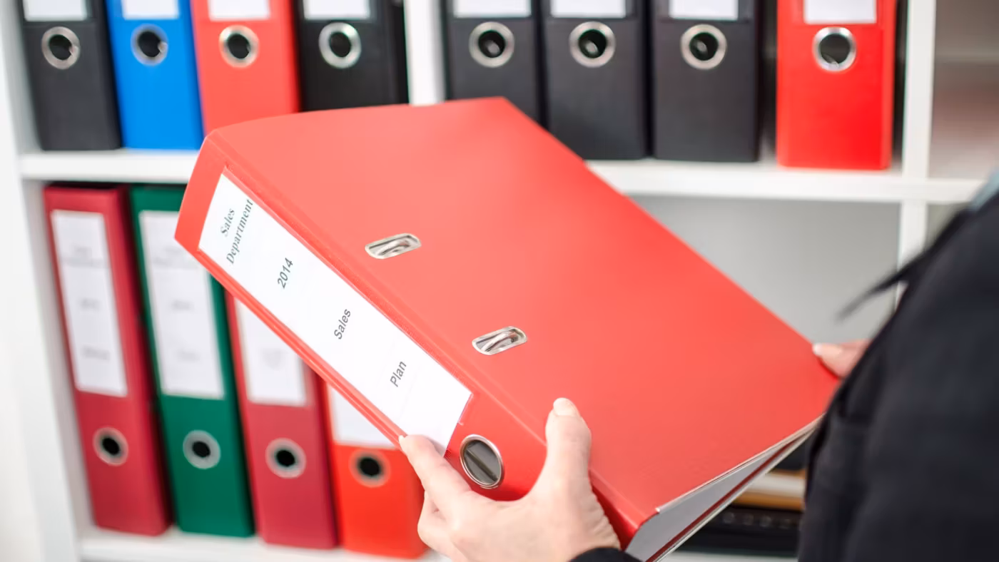 A hand pulling a red lever-arch file from a shelf of archive boxes