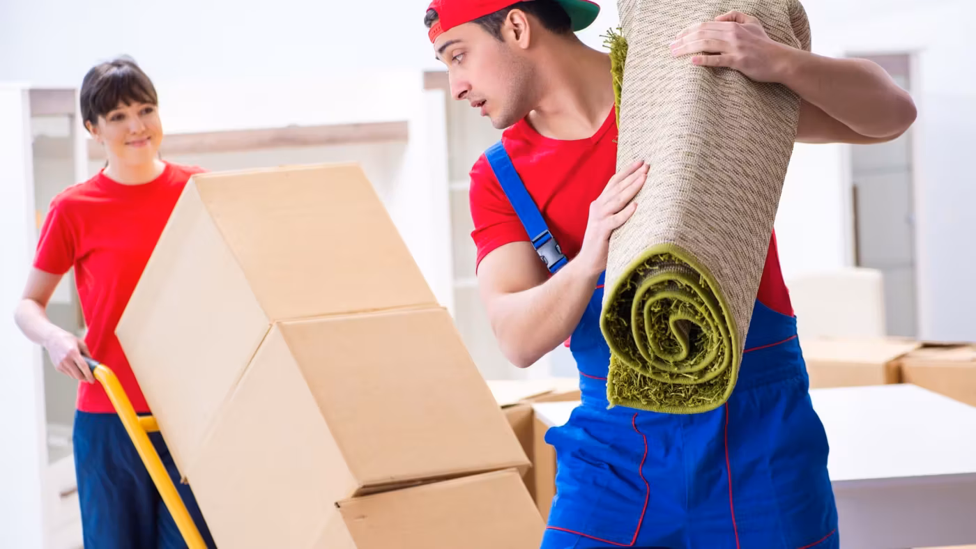 A mover with a rolled rug on his shoulder beside stacked cartons