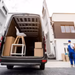The open rear of a van holding a chair and cartons, two movers working beside it with houses behind