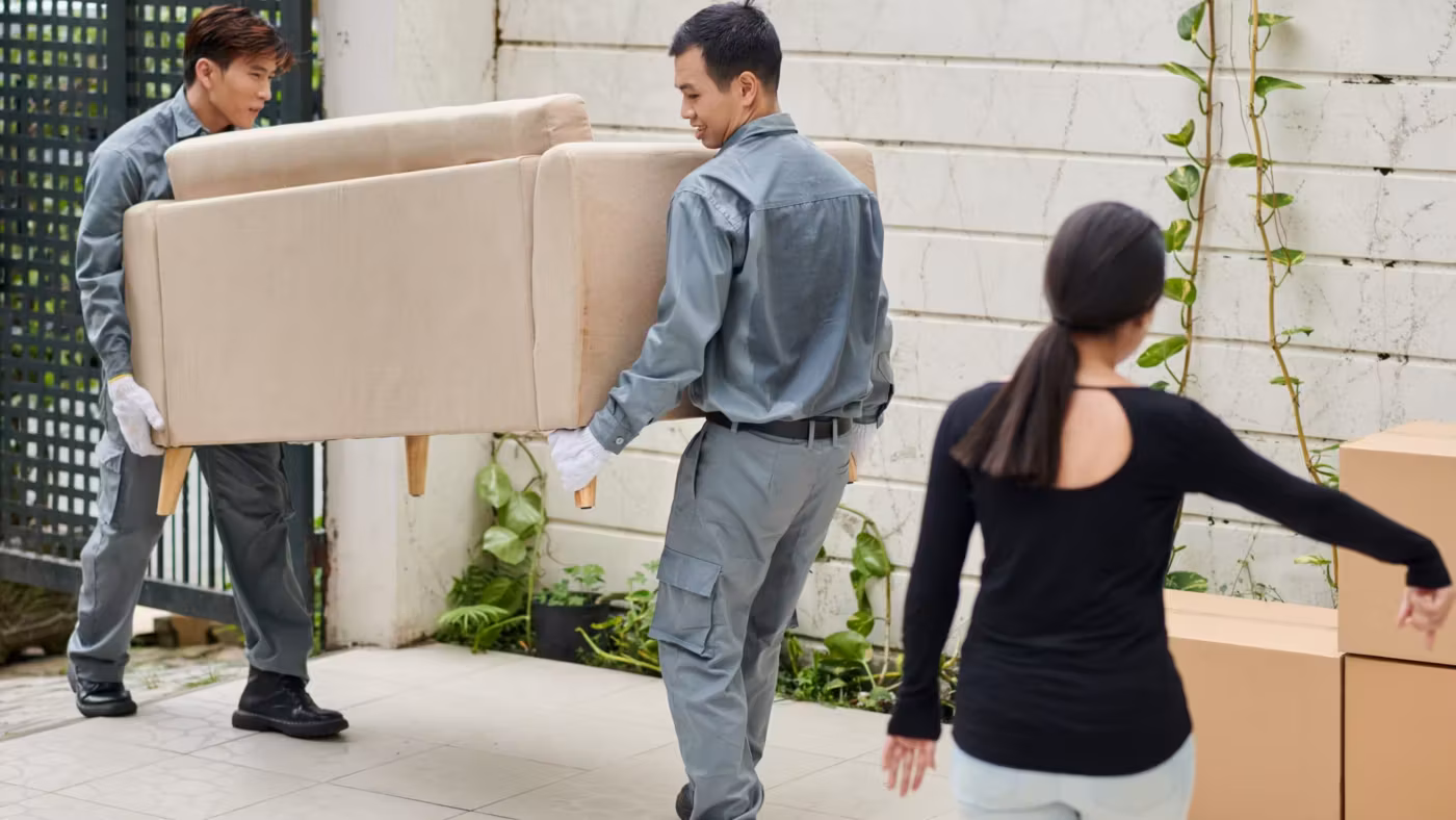 Two movers carrying a cream armchair along a path past a resident, cartons stacked against the wall