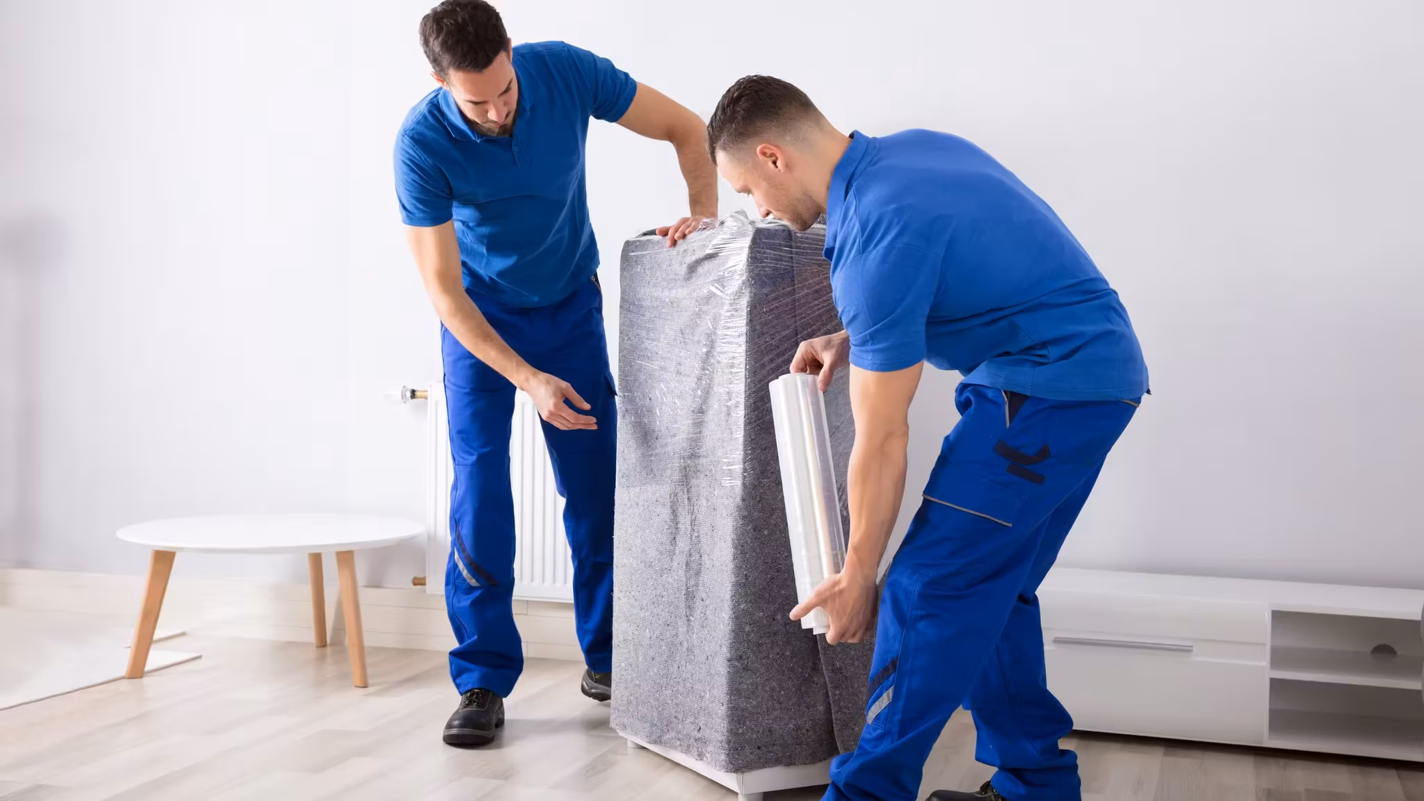 Two movers wrapping a rolled mattress in stretch film in an empty room