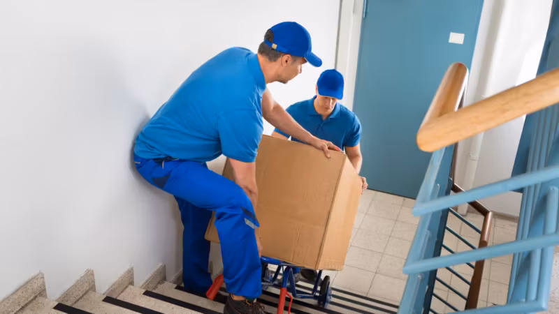 A mover easing a carton on a hand truck up an interior staircase while a second guides it