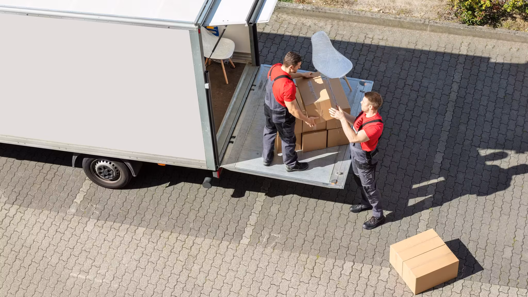 Seen from above, two movers carrying furniture from a box truck across a paved drive