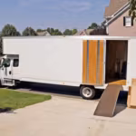 A white moving truck parked at a suburban house with its rear door up and a wooden ramp down to the drive