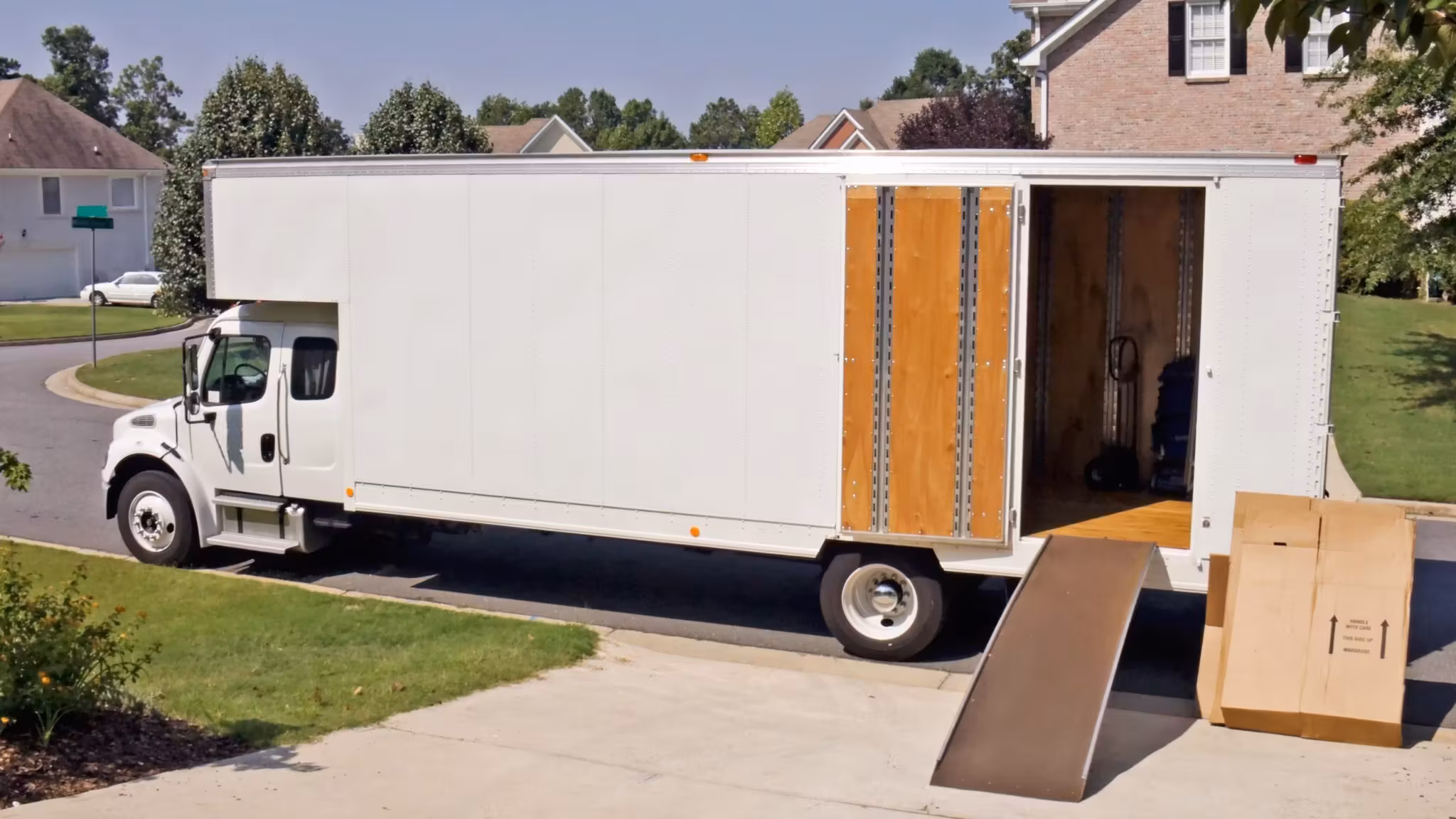 A white moving truck parked at a suburban house with its rear door up and a wooden ramp down to the drive