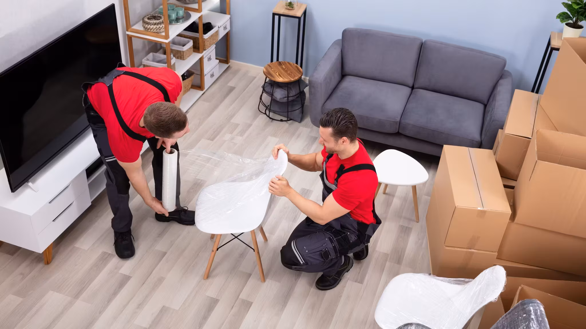 Two movers stretch-wrapping a white chair in a living room stacked with cartons