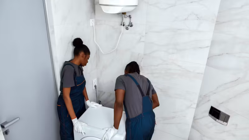 Two movers easing a washing machine away from the wall of a tiled bathroom