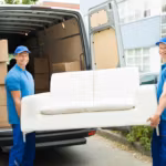 Two movers lifting a white sofa into the back of a van on a residential street