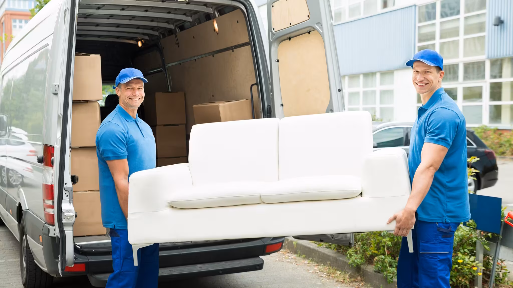 Two movers lifting a white sofa into the back of a van on a residential street