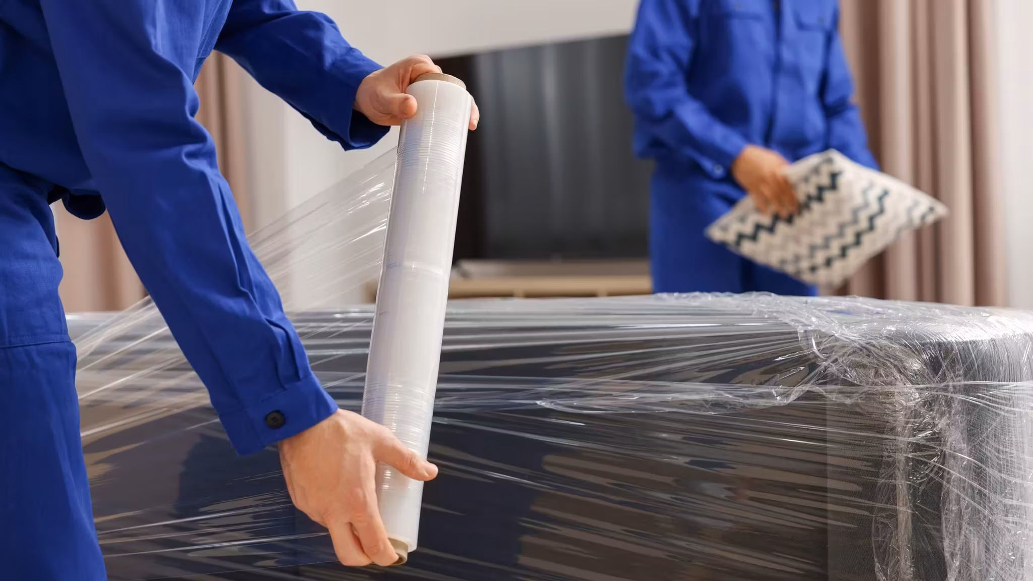 A mover drawing stretch film over a large wrapped item, a second behind him holding folded fabric