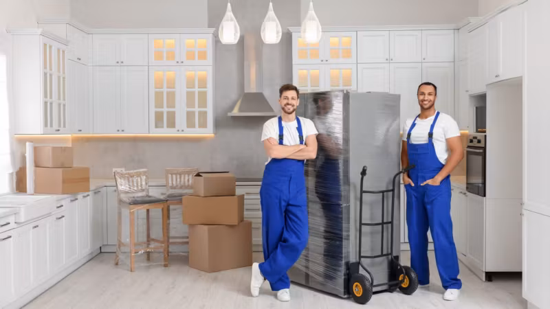 Two movers with a refrigerator strapped to a hand truck in an empty kitchen