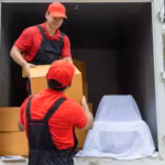 Two movers loading cartons into a box truck alongside a large item under a white cover