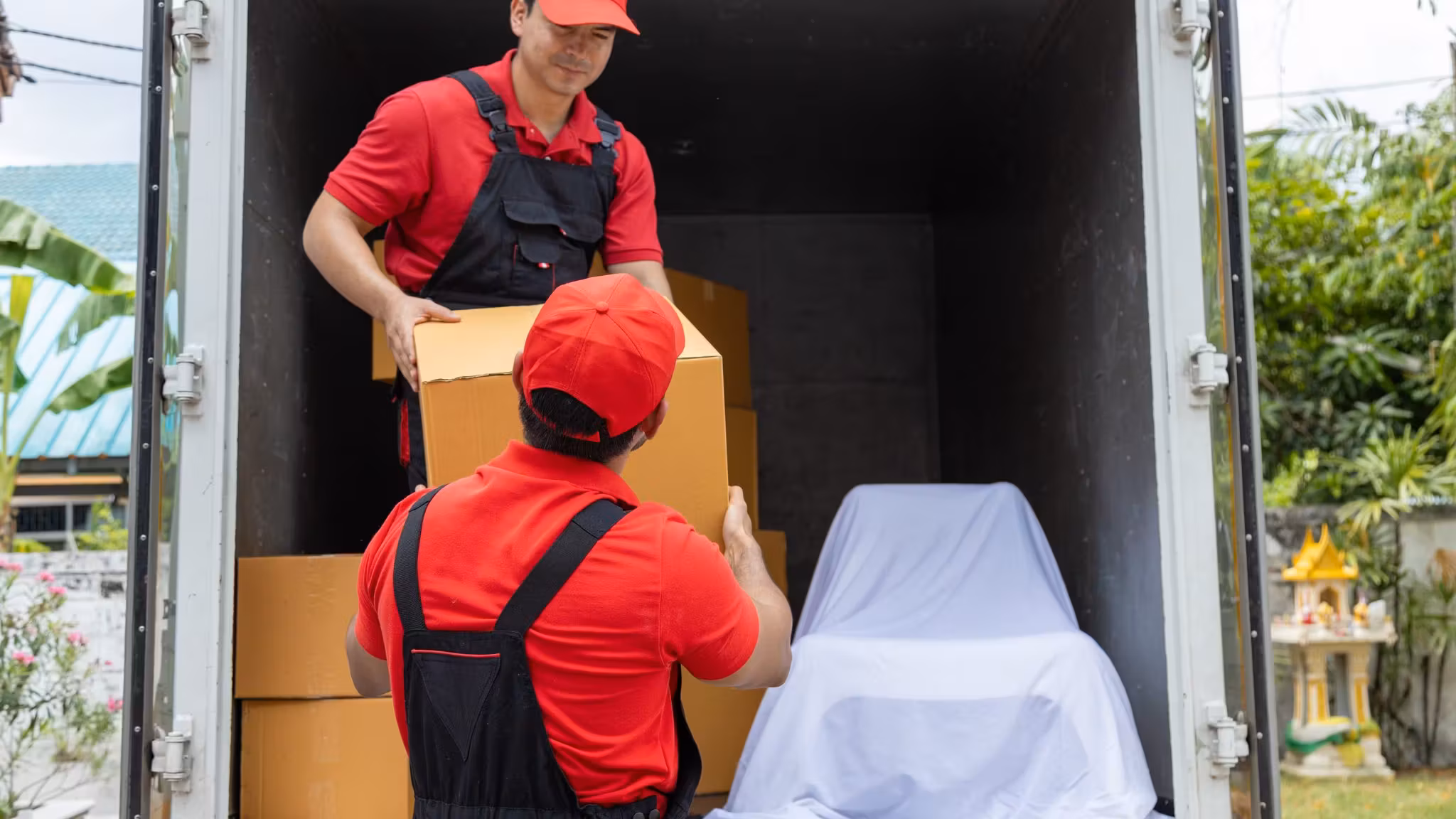 Two movers loading cartons into a box truck alongside a large item under a white cover