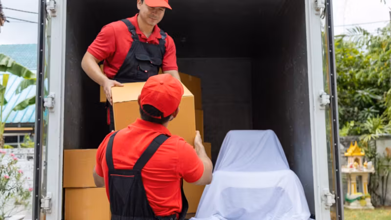 Two movers loading cartons into a box truck alongside a large item under a white cover