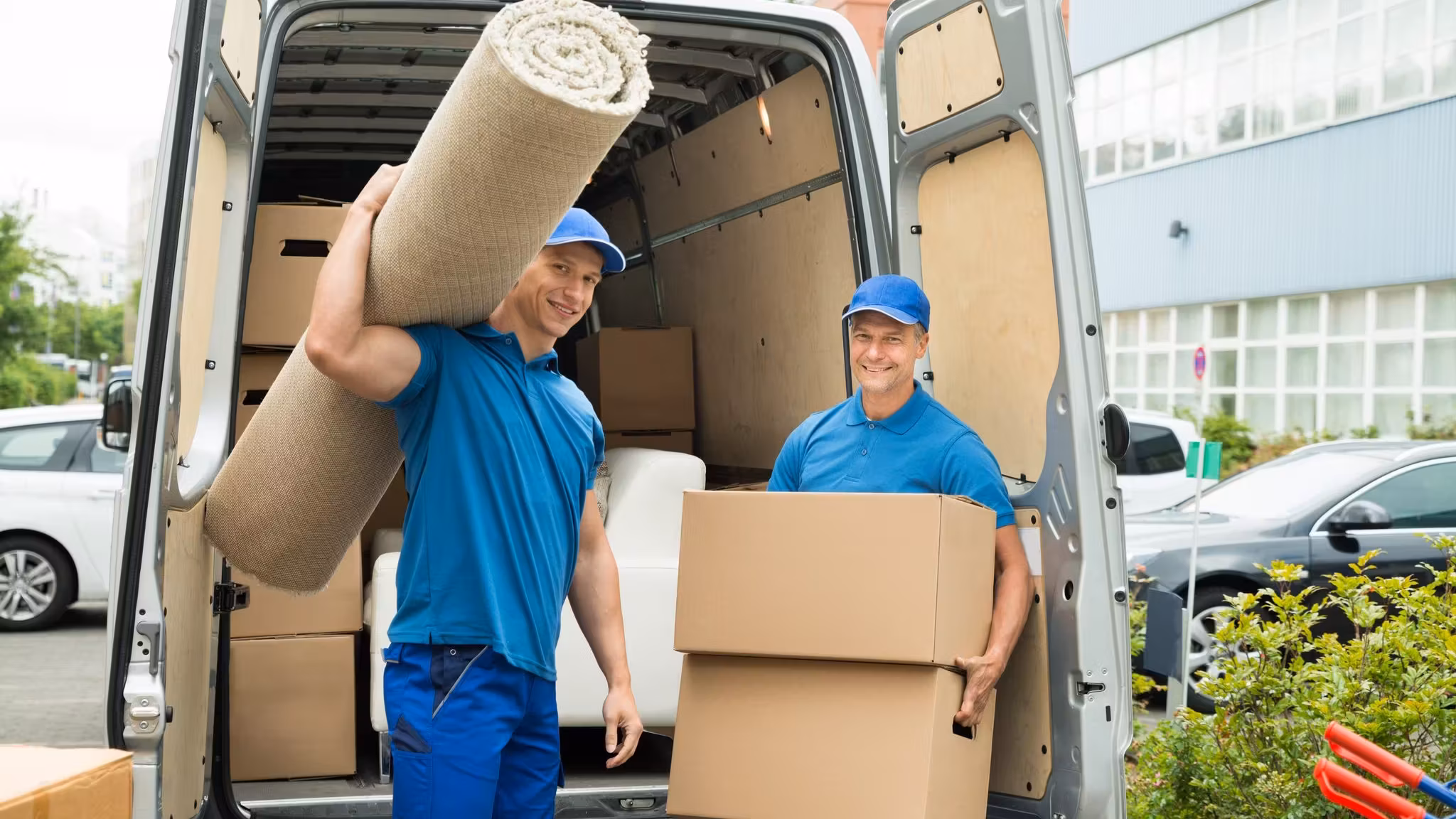 A mover with a rolled rug on his shoulder beside an open van, a second holding a carton