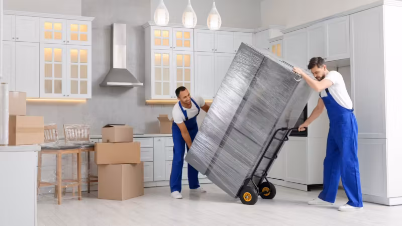 Two movers manoeuvring a wrapped appliance on a hand truck through a kitchen
