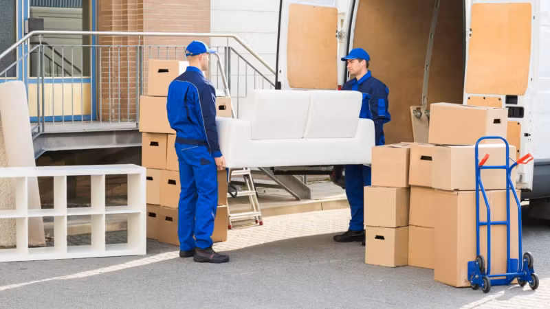 A sofa and stacked cartons on the pavement beside a van, two movers working between them