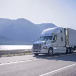 A tractor-trailer on a highway with mountains and open water behind it