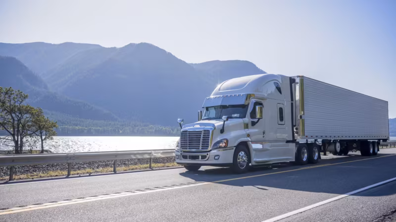 A tractor-trailer on a highway with mountains and open water behind it