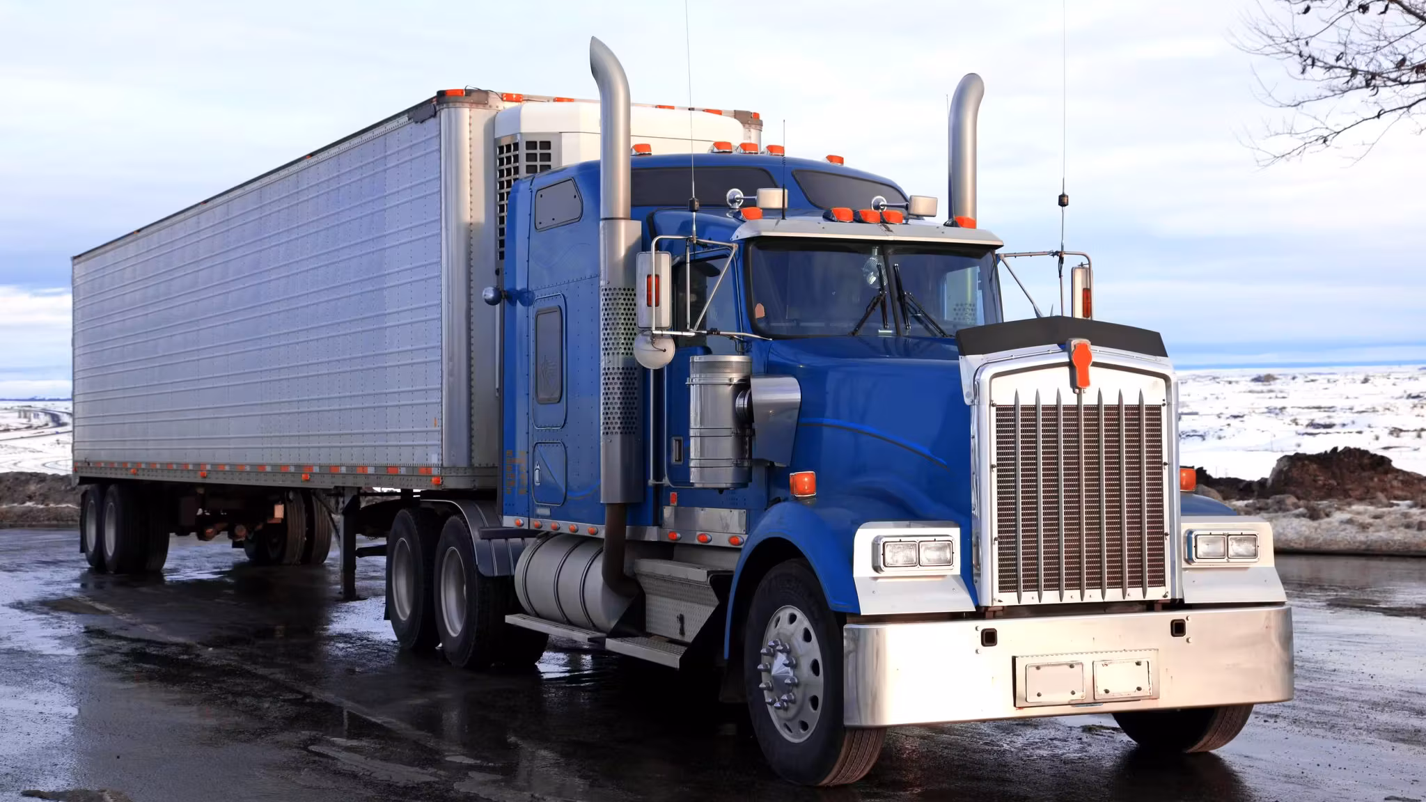 A conventional tractor and dry-van trailer standing on wet ground with snow banked behind