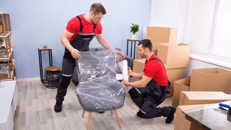 Two movers stretch-wrapping an upholstered chair in a living room stacked with cartons