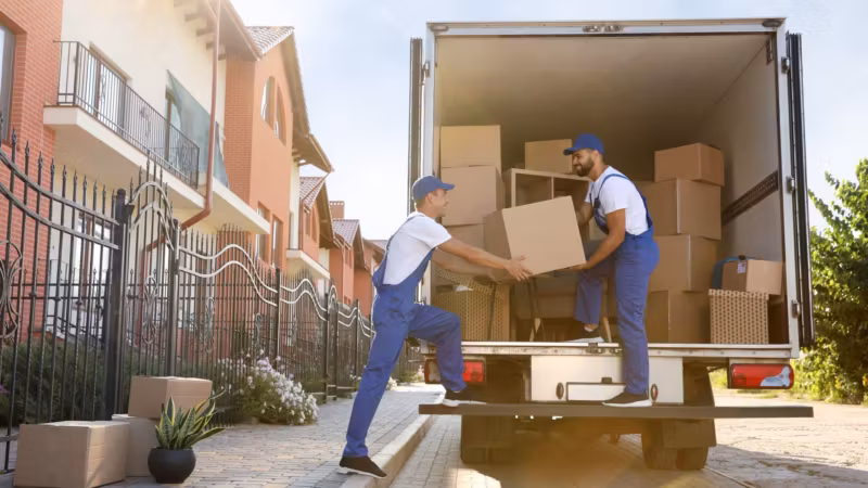 Two movers passing a carton at the open tail of a loaded box truck on a residential street