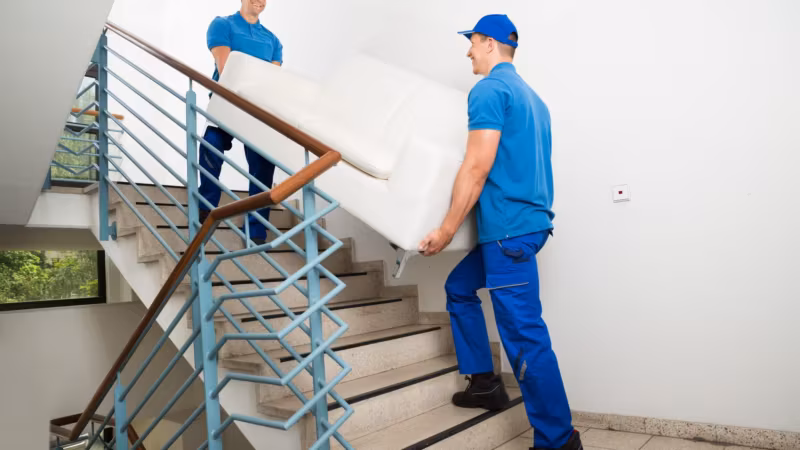Two movers carrying a white sofa up an interior staircase