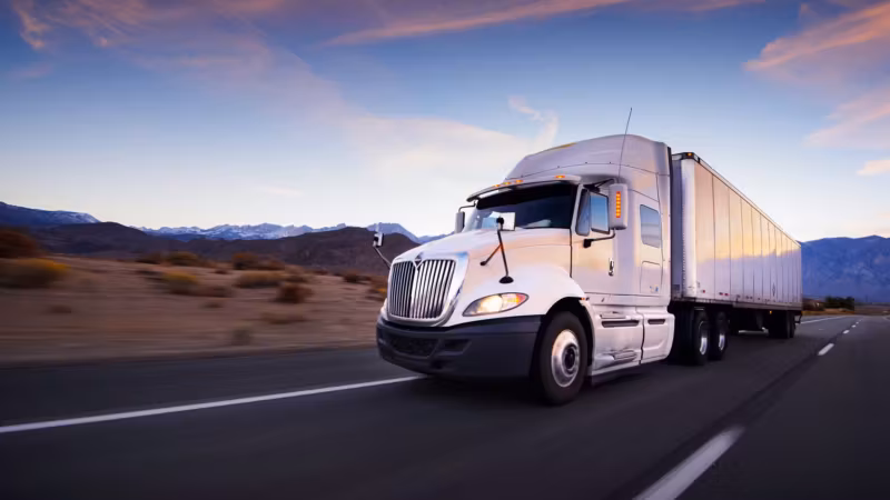 A white tractor-trailer traveling an open desert highway at dusk, mountains in the distance