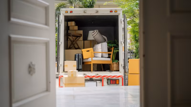 A loaded moving truck seen through the part-open front door of a house, chairs and cartons stacked inside it