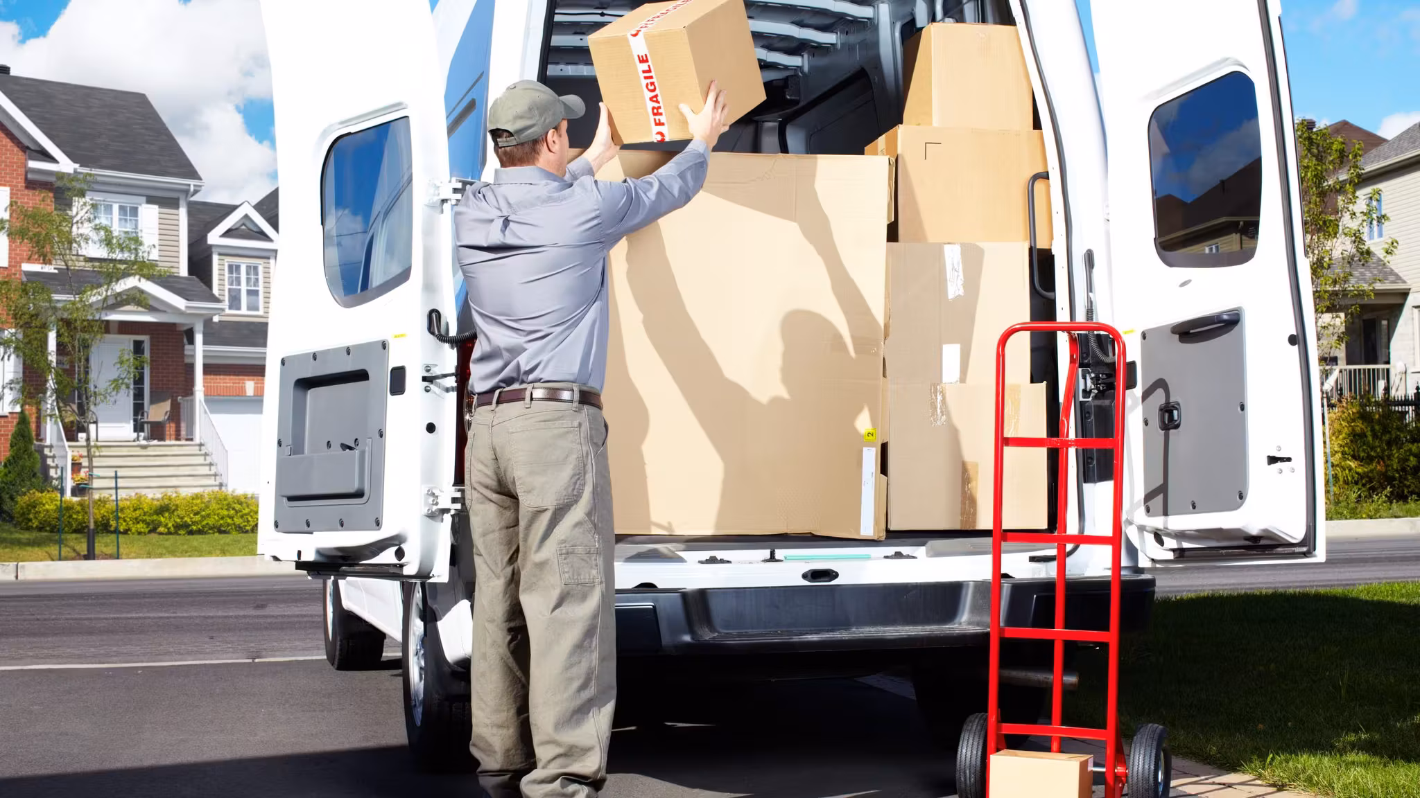 A mover stacking cartons into the back of a van at the curb, a red hand truck beside him and houses behind