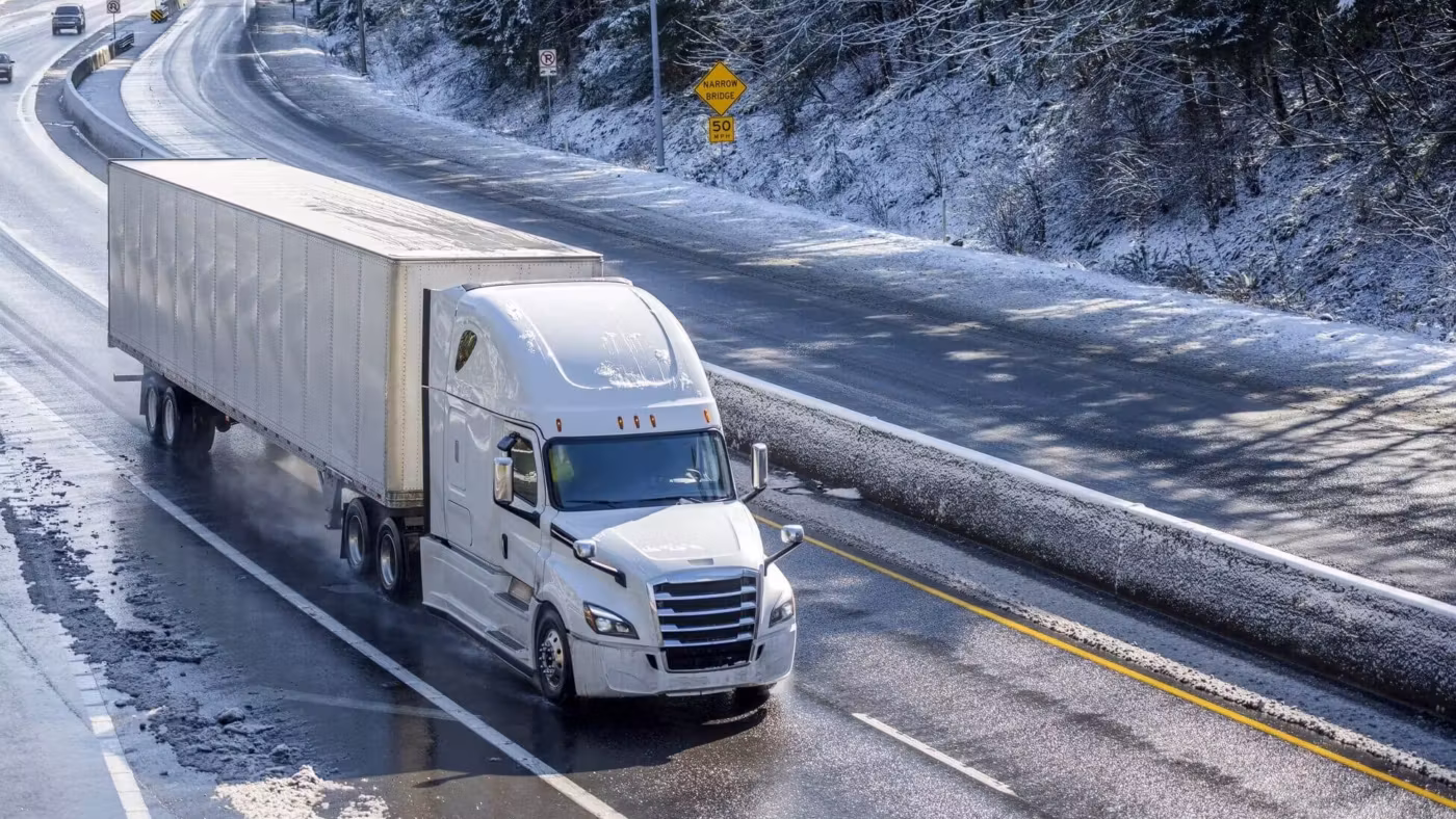 A semi-trailer on a wet highway curve between snow-covered banks, traffic ahead