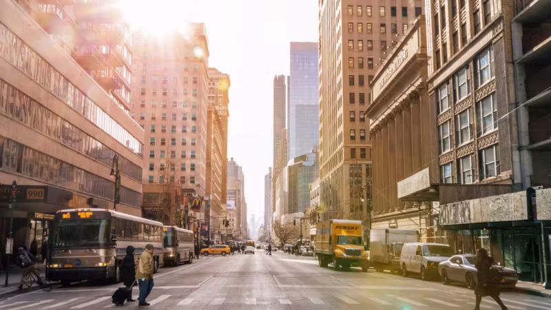A Manhattan avenue at sunrise with a bus, a cyclist and a traveller pulling a suitcase
