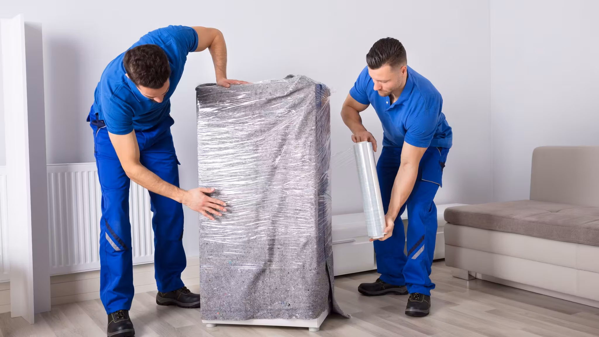 Two movers wrapping a tall cabinet in protective film before it travels