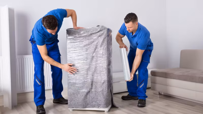 Two movers wrapping a tall cabinet in protective film before it travels