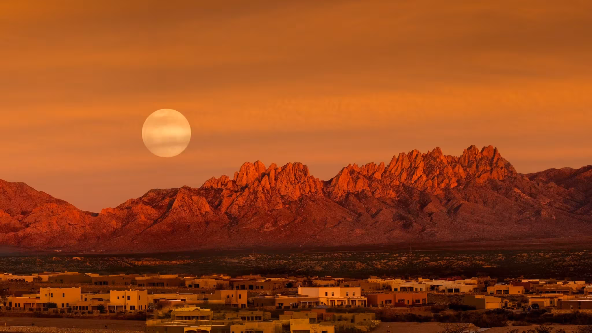 A full moon above red mountains at sunset with adobe houses on the plain below